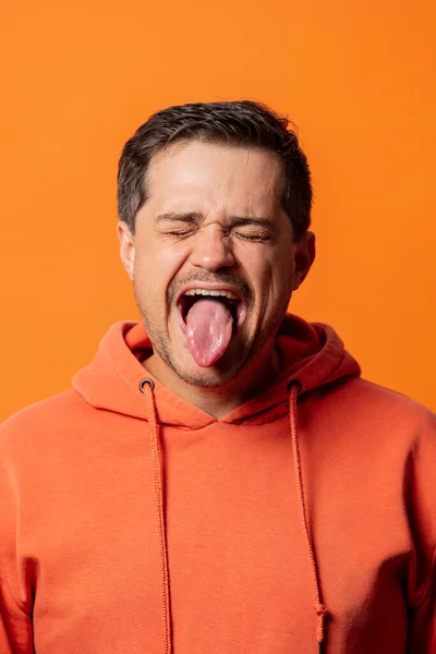 オレンジ色のパーカーの男はオレンジの背景に舌を見せ — ストック写真