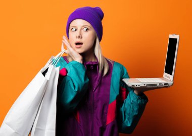 90 'ların eşofmanlı, turuncu arka planda not defteri ve alışveriş torbalarıyla stil sahibi kadın.