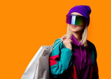 90 'ların eşofmanlı stil kadını ve turuncu arka planda alışveriş torbaları olan VR gözlükleri.