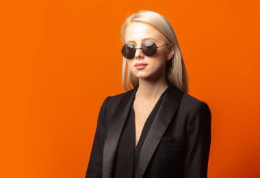 Siyah ceketli ve turuncu arka planda güneş gözlüklü şık bir sarışın.
