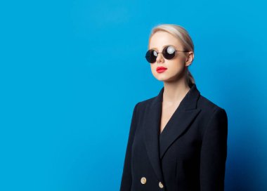 Siyah ceketli şık bir sarışın ve mavi arka planda güneş gözlüğü.