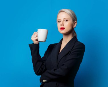 Mavi arka planda fincanı olan siyah ceketli şık bir sarışın.