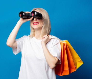 Alışveriş torbaları ve mavi arka planda dürbünlü sarışın. 