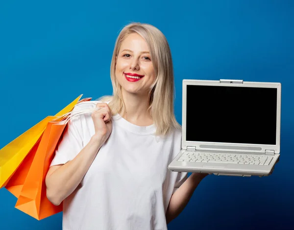 穿着白色T恤 蓝色背景的购物袋和笔记本电脑的金发美女 — 图库照片