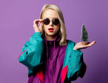 80 'lerin rüzgarlığı ve mor arka planda Noel ağacı olan yuvarlak güneş gözlüklü tarz sarışın.