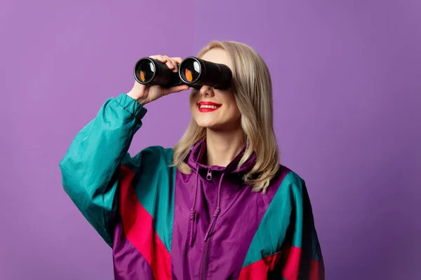 80年代のスタイルの女性ウインドブレーカーとラウンドサングラスは紫の背景に双眼鏡を保持 — ストック写真