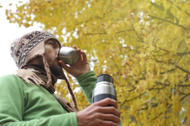 yün örgü şapkalı sakallı adam kupa, sarı sonbahar yeşillik zemin üzerine alt yan görünümü üzerinden sıcak çay veya kahve içer