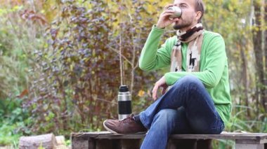 örme eşarp oturan sakallı hipster ve dinlenme bacaklarını geçti, içecekler - fincan çay veya kahve ve doğa güzelliği zevk, termos yakın ve terk etmek üzere, yan görünüm, arka plan sonbaharda 