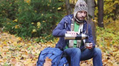 şapka ve eşarp oturan sakallı hipster adam oturma ve sonbahar ormanda dinlenme, sıcak içecek döker - çay veya kahve termos fincan içine, kupa üfleme ve içecekler, rüyalar ve doğa güzelliği zevk 
