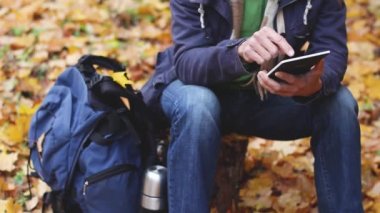 bin yıllık adam blogger kütük ve çalışma üzerinde ormanda oturan - tablet bilgisayarda bir blog yazıyor, ya da ona yakın harita, sırt çantası ve termos görünümlü, yürüyüşten sonra dinlenme, sonbahar yaprakları arka planda 