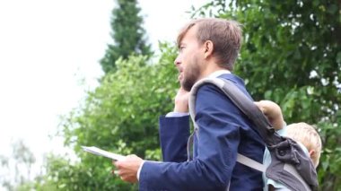 akıllı telefon üzerinde belgeler le mavi takım elbiseli sakallı bin yıllık işadamı, sinirli sözleşme tartışıyor, ağlayan oğlu sakinleştirmeye çalışıyor, sırtında askıda bebek çocuk, açık yeşil yaz yaprakları 