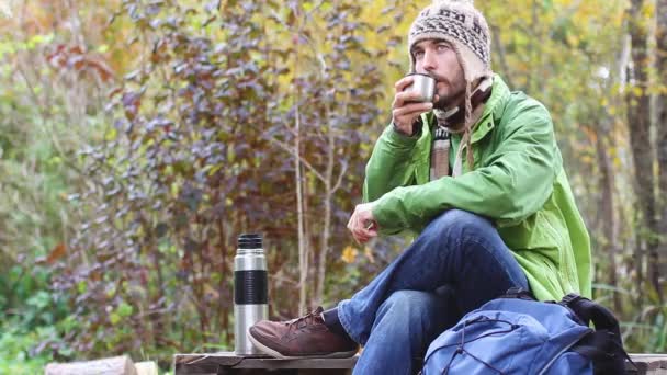 时髦的胡须千禧年男子在针织帽子坐在和休息交叉他的腿 喝热茶或咖啡从杯子和梦想 背包和热水瓶在他附近 野餐在秋天的森林 — 图库视频影像
