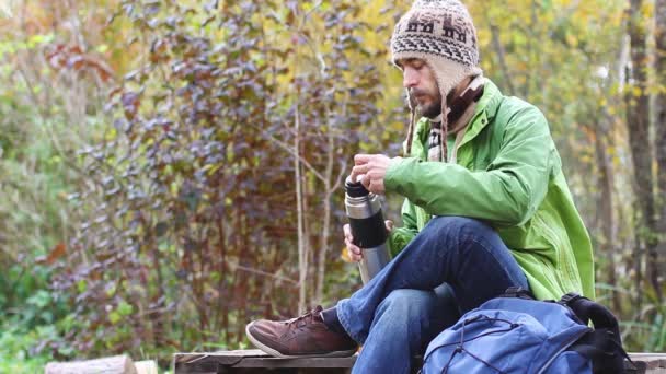 孤独的时髦旅行者男子在针织帽子与胡子坐和休息交叉他的腿 倒热饮 茶或咖啡从热水瓶到杯子 吹在杯子和饮料 背景秋天森林 — 图库视频影像