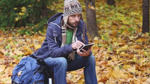 千禧年留着胡须的时髦博主 戴着针织帽子和围巾坐在树桩上的森林里 在平板电脑上写博客 看地图 背包靠近他 徒步旅行后休息 秋天的树叶 — 图库视频影像