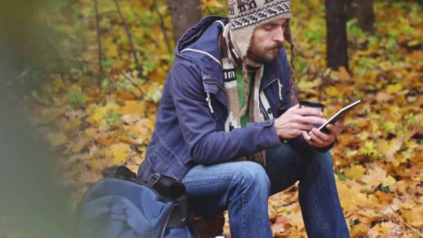 千禧年留着胡须的时髦博主 戴着针织帽子和围巾坐在森林里 在树桩上 喝着热茶或咖啡 在平板电脑上工作 在他身边的背包 在秋天的树叶背景 — 图库视频影像