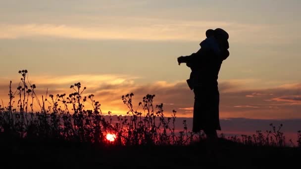 母亲与婴儿在怀里转过身来 亲吻他 并显示他的日落 剪影在日落背景在外地 — 图库视频影像