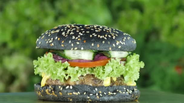 Hamburger of hamburger met een zwarte broodje en sesamzaden op onscherpe achtergrond van greens en groenten. — Stockvideo