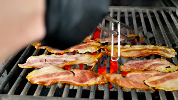 Baconskivor är stekt på en grill, jag vrider bacon en efter en med järn tång till en annan för att få den till beredskap — Stockvideo