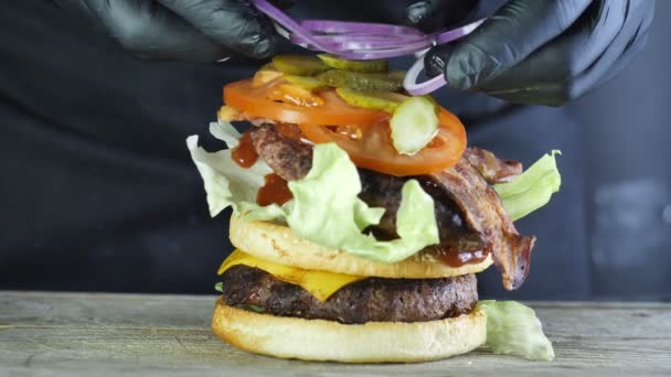 Mega hamburguesa grande con una chuleta doble con verduras y un bollo de sésamo, el proceso de formar una hamburguesa, el chef en guantes negros pone rodajas de cebolla en la capa superior de la hamburguesa — Vídeo de stock