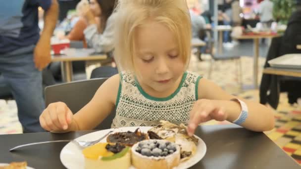 Bebé niña intenta surtidos pasteles en un café sobre un fondo borroso — Vídeos de Stock