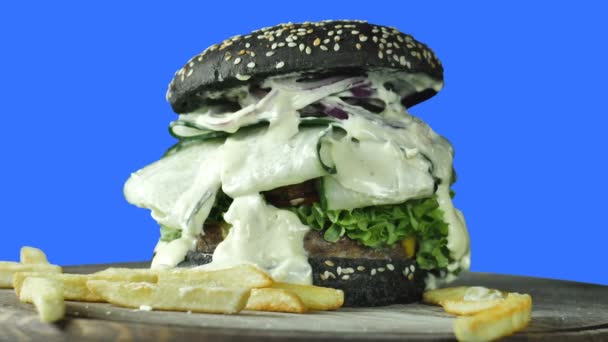 채소와 감자 튀김으로 둘러싸인 고기 패티와 함께 녹색 빵을 뿌린 큰 육즙이 많은 햄버거는 고립 된 파란색 배경, 패스트 푸드에서 회전합니다. — 비디오