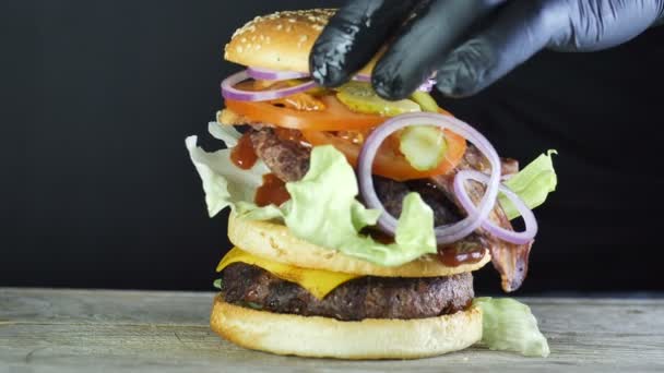 Mega Big Burger mit doppeltem Schnitzel mit Gemüse und Sesam-Brötchen, der Prozess der Bildung eines Burgers, der Koch in schwarzen Handschuhen breitet auf der obersten Schicht des Burgers die letzte Zutat Hälfte der oberen — Stockvideo