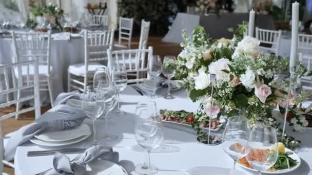 다양한 맛있는 요리를 위한 흰색 테이블보가 있는 대형 테이블이 있는 연회, 결혼식 이나 생일을 위한 케이터링 서비스가 있는 장미 양초 장식이 있는 꽃꽂이. 카메라 이동 속도가 느립니다. — 비디오