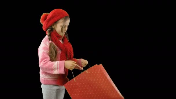 Een meisje van ongeveer tien in een rode gebreide muts en sjaal met Pigtails en met een pakket in haar handen opent en kijkt in een pakket met geschenken en verbaast een langverwachte gift, winter schieten op een — Stockvideo
