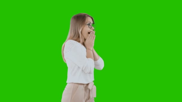 A young blonde girl wearing white blouse and pants looks to the side and emotionally admires bringing hands to her mouth, holidays, green background — Stock Video