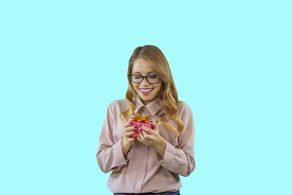 Young beautiful girl holds a small box with a bow looks at her and smiles on a blue isolated background — Stock Photo, Image