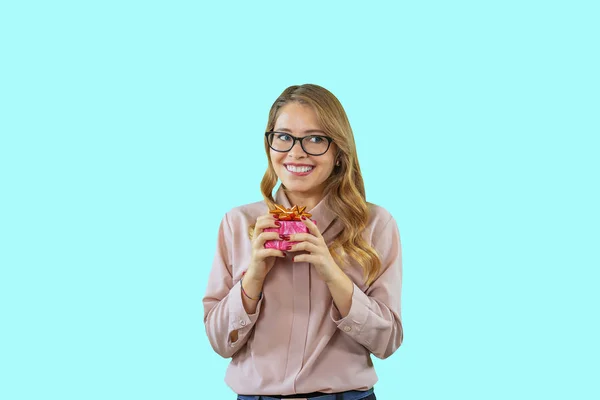 Ένα χαριτωμένο νεαρό κορίτσι με γυαλιά και μια ροζ μπλούζα κρατά ένα δώρο, ενώ χαμογελά, ενώ περιμένει και κοιτάζοντας πλάγια, δαγκώνοντας τα χείλη της σε ένα μπλε απομονωμένο φόντο — Φωτογραφία Αρχείου