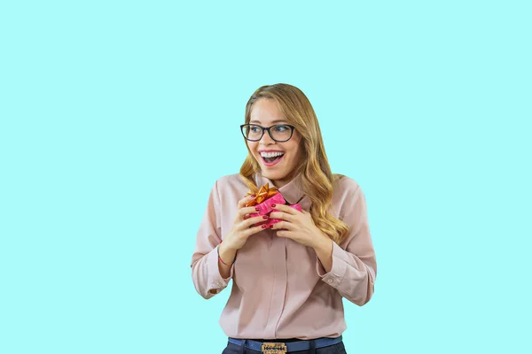 Ein schönes junges Mädchen in Brille und rosa Bluse hält ein Geschenk aufrichtig erfreut und freudig zur Seite blickend auf einem blau isolierten Hintergrund — Stockfoto