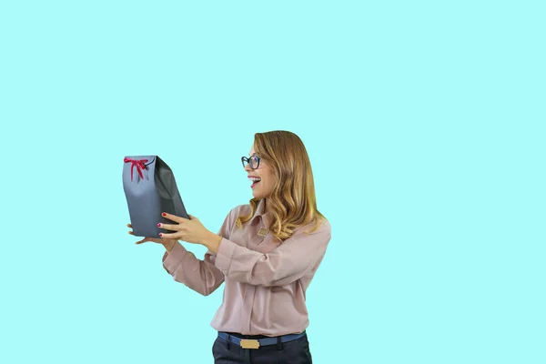 Portrait of a young beautiful blonde in glasses holding a box with a red velvet ribbon on her outstretched hands and looking sideways smiling on an isolated background, New Year holidays, birthday — Stock Photo, Image