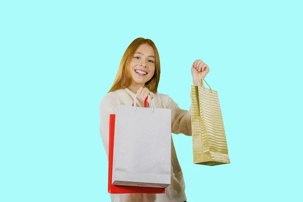 Aantrekkelijk meisje met lang rood haar glimlacht, kijkt naar de camera en houdt papieren tassen met cadeaus op een geïsoleerde blauwe achtergrond — Stockfoto