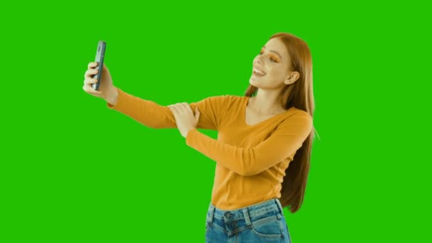 Молодая девушка с длинными волосами делает различные позы и улыбается на камеру телефона, селфи, зеленый фон — стоковое видео