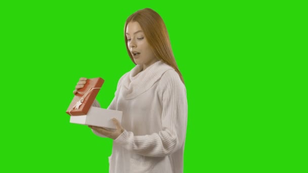 Attractive and young girl with long red hair in a white sweater opens gift on an isolated background — Stock Video