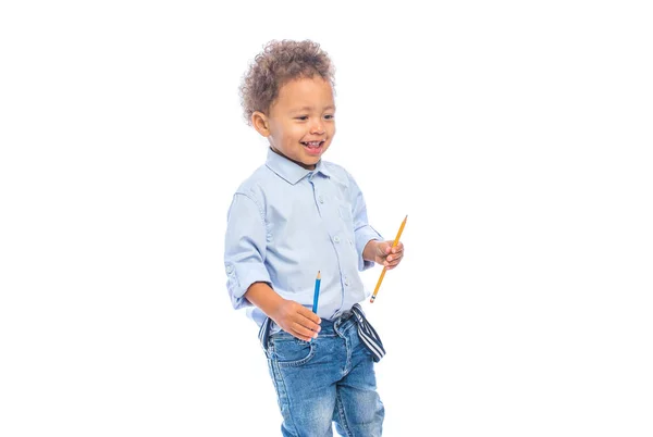 Ein Porträt eines kleinen dunkelhäutigen Jungen mit lockigem Haar in Jeans und hellem Hemd steht im Profil und hält verspielt Bleistifte — Stockfoto