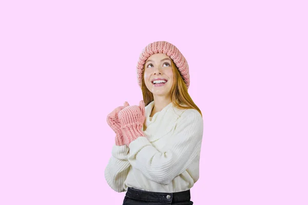 Attractive red-haired girl in a warm hat and mittens looking at the camera against an isolated pink background — Stock Photo, Image