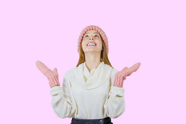 Young cute red-haired teen girl in a knitted pink hat and mittens holding her hands up looks up and smiles on an isolated pink background, girls emotions, winter holidays and holidays — 스톡 사진