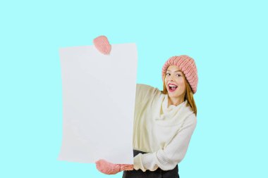Elinde poster olan bir kız. Örme pembe şapkalı genç kızıl saçlı bir kız, iki eliyle de yan yana bir poster tutuyor ve izole bir arka plan üzerinde yan bakarak gülümsüyor