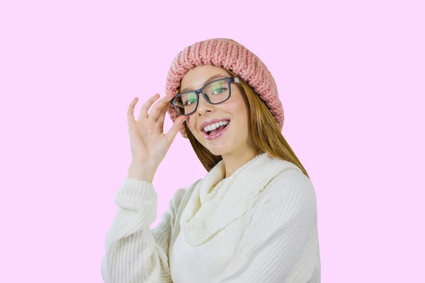 Молодая девушка с рыжими длинными волосами в розовой вязаной шляпе и в очках стоит в половине оборота держа очки улыбаясь с открытым ртом и глядя на камеру на изолированном фоне — стоковое фото