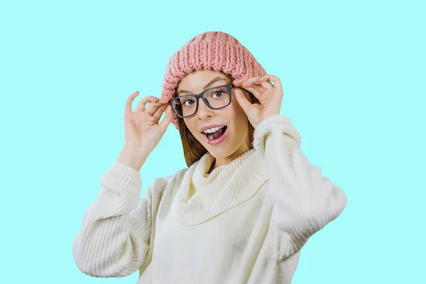 A young girl with red long hair in a pink knitted hat and in glasses stands half turn holding glasses with both hands enthusiastically smiling with her mouth open and looking at the camera against an — Stock Photo, Image