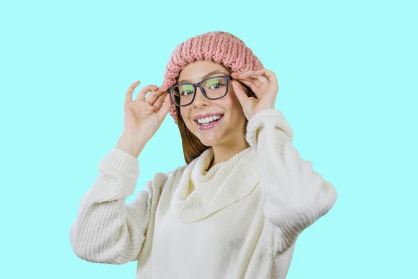 분홍색 니트 모자와 안경을 쓴 빨간 긴 머리를 한 어린 소녀가 두 손으로 미소를 지으며 고립된 배경에서 카메라를 바라보며 안경을 들고 반쯤 서 있습니다. — 스톡 사진