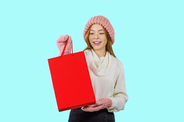 Portrait of a cute red-haired girl in a knitted hat and mittens holding a red bag and looking at it showing something on an isolated background, fashion shooting, New Years gifts — Stock Photo, Image