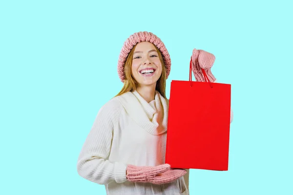 Retrato de uma menina bonita ruiva em um chapéu de malha e luvas segurando um saco vermelho e olhando para a câmera e rindo mostrando um presente em um fundo isolado, tiro de moda, presentes de Ano Novo — Fotografia de Stock