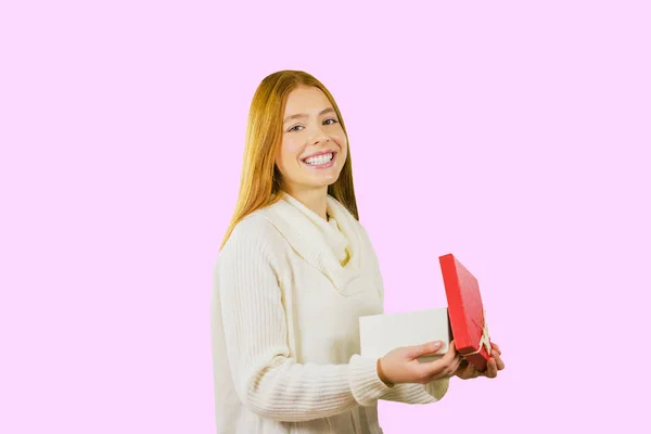 Ελκυστικό κορίτσι στέκεται στο προφίλ με ένα ανοιχτό κουτί με ένα δώρο χαμόγελα και κοιτάζει μέσα στην κάμερα, απομονωμένο φόντο — Φωτογραφία Αρχείου