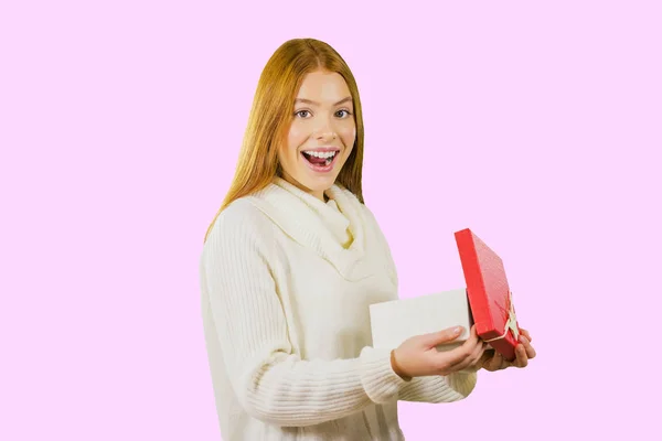 Jolie jeune fille aux cheveux roux debout de profil avec une boîte ouverte avec un cadeau sourit avec une bouche ouverte et regarde dans la caméra, fond isolé — Photo