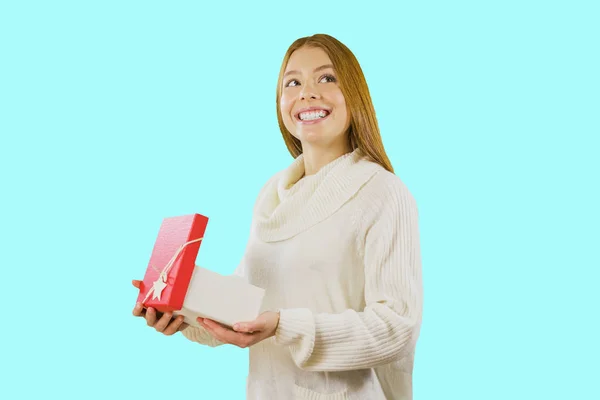 Portré egy aranyos vörös hajú lány áll, félig megfordul, és tart nyitva egy dobozt egy ajándékot, és mosolyog, és úgy néz ki titokzatosan fel az oldalra egy elszigetelt háttérrel. Szilveszteri szünidő, születésnap — Stock Fotó