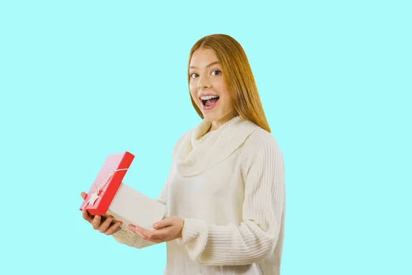 Mulher bonita com cabelo vermelho em uma camisola branca posando com uma caixa de presente. Retrato de uma jovem que segura uma caixa aberta e sorri de surpresa e olha para a câmera, fundo isolado — Fotografia de Stock