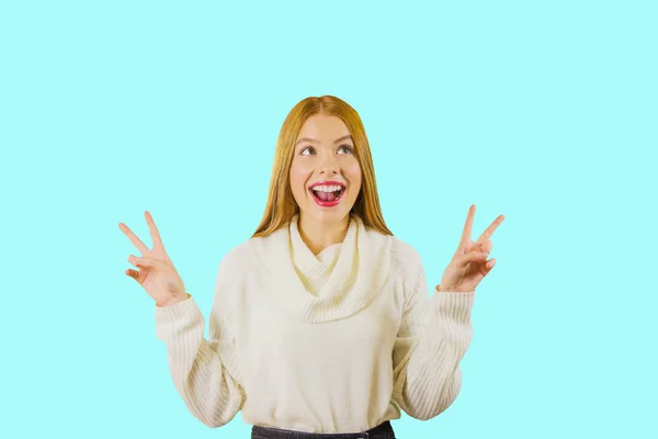 Portrait of a young girl with long red hair showing piss with both hands holding them up, smiling, looking to the side up on an isolated background — Stockfoto